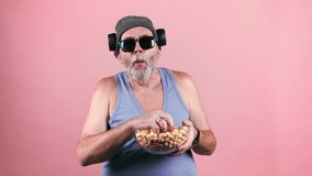 Senior man in blue tank top, sunglasses, grey hat, wireless headphones eagerly eats popcorn in handfuls from glass bowl. Isolate on pink background.