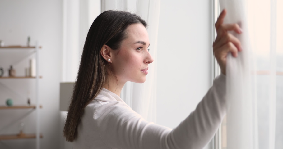 Young woman opening curtain looking through window. Happy confident lady enjoying beautiful view and dreaming at home. Smiling girl contemplating feeling hope peaceful morning standing in apartment Royalty-Free Stock Footage #1048391239