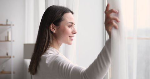 Young woman opening curtain looking through window. Happy confident lady enjoying beautiful view and dreaming at home. Smiling girl contemplating feeling hope peaceful morning standing in apartment