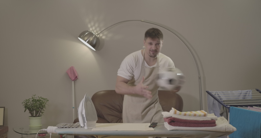 Active young Caucasian man with soccer ball cheering as watching match on TV indoors. Excited handsome guy making victory gesture standing in front of ironing board with laundry. Cinema 4k ProRes HQ.