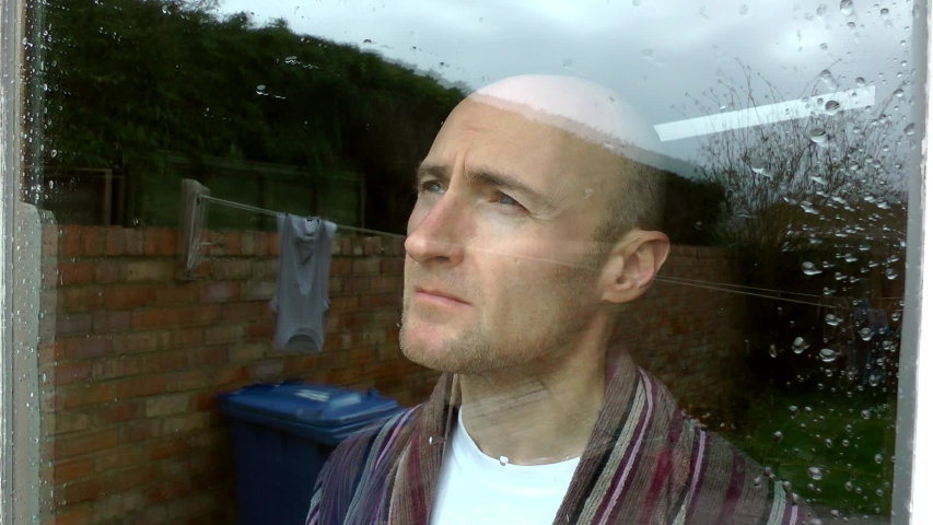 A midlife crisis. Male looks out of house window depressed and bored. Middle aged man wearing a dressing gown. Mental health, self isolation midlife crisis, financial dept.