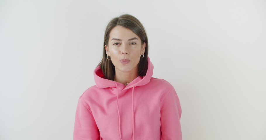 Portrait of young sexy playful girl dressed in pink hoodie standing over white background blowing bubblegum candy bubbles and chewing gum. Woman looking at camera. 4k raw video footage slow motion