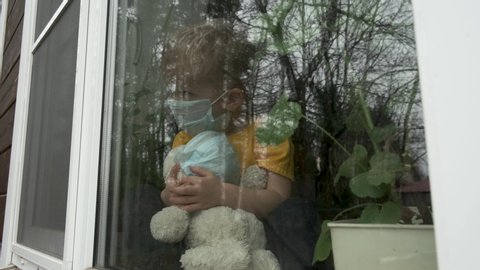 Stay at home quarantine for coronavirus pandemic prevention. Child and his teddy bear both in protective medical masks sits on windowsill and looks out window. View from street. Prevention epidemic