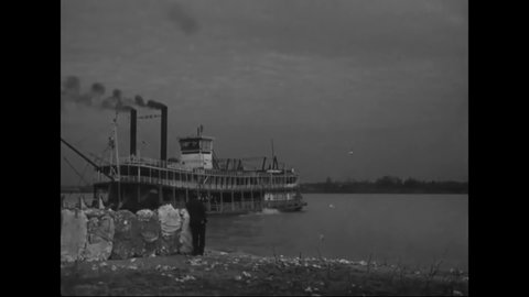 CIRCA 1930s - Pickers stand with bales off cotton on the shoreline as an old paddle steamer passes by on the Mississippi river.