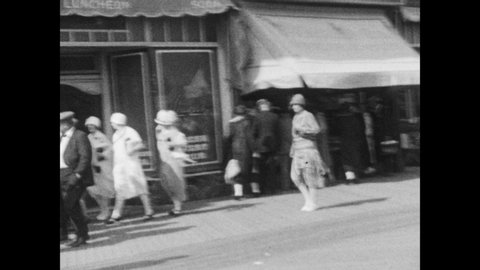 1920s: crowd walks in front of stores and bathhouse, 2 women walk with arms around each other, man pushes cart with umbrella and women, boy walks by and looks at camera, person with umbrella walks by