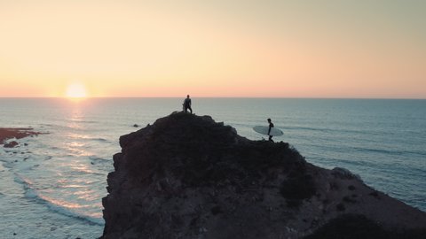 Amazing aerial shot of woman and man surfers standing at the edge of cliff / rock and watching waves at sunrise / sunset. Use for fitness, lifestyle advertising, commercial. : vidéo de stock