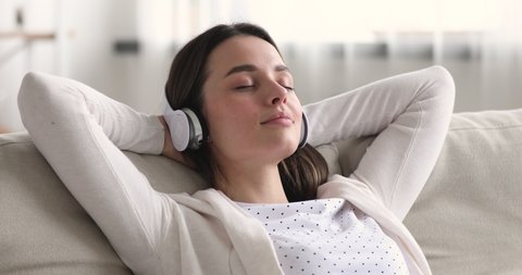 Calm relaxed young woman chilling on couch with eyes closed wearing headphones. Serene happy millennial girl listening cool music or modern audio book lounge at home holding hands behind head.