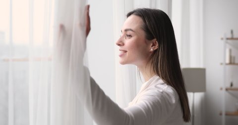 Happy young woman opens window lace looking outside apartment. Smiling mindful attractive lady watching beautiful cityscape view enjoying dreaming and contemplating feeling hope concept. Side view