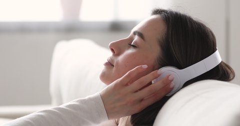 Serene young woman relaxing on comfortable sofa with eyes closed wearing headphones. Pretty lady enjoys listening chill music audio sound meditating feeling no stress at home. Close up side view
