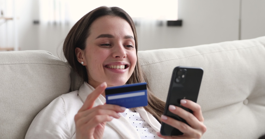 Happy young woman holding credit card using instant mobile payments at home. Smiling beautiful girl customer shopper making purchase on smart phone receiving cashback concept. E-banking app service