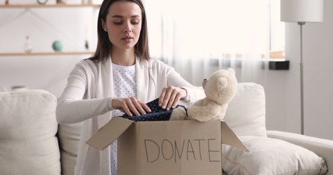 Beautiful young woman packing clothes, toy in children donation cardboard box concept. Female volunteer looking at camera pointing finger inviting you to participate in voluntary work and help kids.