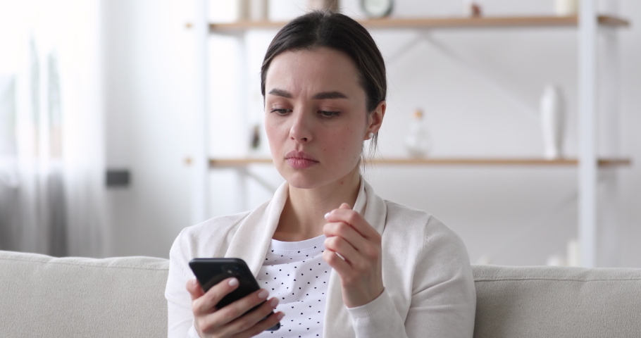 Worried nervous young woman reading bad news in message on smartphone at home. Stressed anxious lady feeling jealous upset holding mobile phone thinking of problem receiving sms sitting alone on sofa. Royalty-Free Stock Footage #1048429339