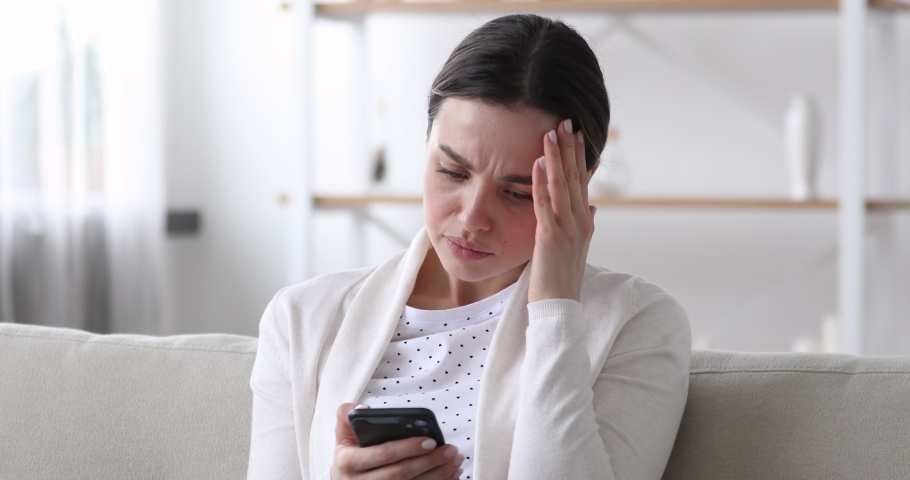 Worried nervous young woman reading bad news in message on smartphone at home. Stressed anxious lady feeling jealous upset holding mobile phone thinking of problem receiving sms sitting alone on sofa. | Shutterstock HD Video #1048429339