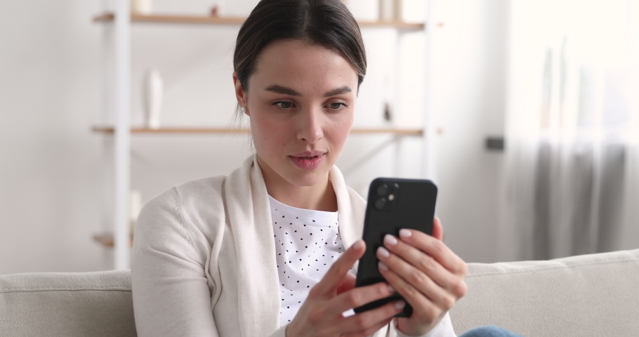 Beautiful young woman looking at smartphone surfing social media dating apps. Pretty millennial girl watching video stories, checking feed, reading news ot texting messages using mobile phone at home. Royalty-Free Stock Footage #1048429342