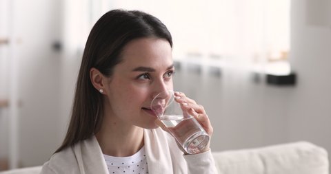 Attractive healthy millennial woman holding glass drinking fresh mineral water at home. Thirsty young lady hydrating thirst tasting pure water in the morning for diet and weight loss balance concept.
