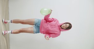 Young woman blowing balloon over white wall background . Girl in pink hoodie dancing and having fun at home or studio. Vertical 4k raw video footage slow motion