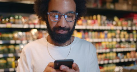 Close up of man using his smartphone while standing in food store. Bearded guy in glasses texting with wife on mobilephone while doing shopping in supermarket. Blurred background.