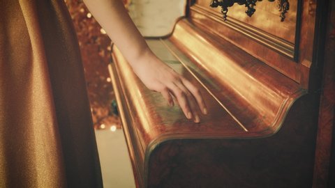 film style 1920s. Closeup beautiful female hand elegantly gently touches with finger old brown retro piano on backdrop bright luminous gold shiny wall. luxurious vintage dress. Cropped face