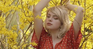 Tender portrait of the charming blonde girl posing among branches of yellow flowers. Young attractive woman with a magnificent golden hair is chilling on the nature. 4K video.