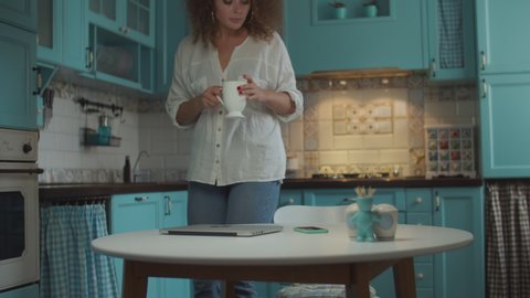 Young curly 20s woman making tea and coming to table with cup. Smiling female opening laptop for working at home on blue kitchen. 