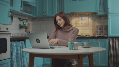 Young 30s woman talking by mobile phone emotionally with hands gesticulating, typing on laptop, sitting at the table on home blue kitchen. 