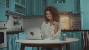 Young curly 20s woman has video call on laptop on kitchen. Female talking emotionally to laptop screen with hands gesticulating, sitting at table at home