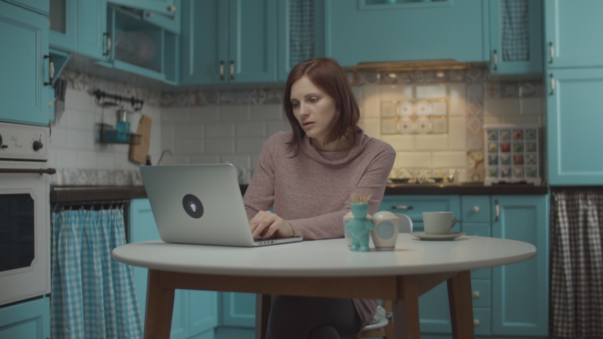 Young 30s female tired of work at home, closing laptop and taking a cup of coffee. Woman making working pause to enjoy hot drink at home blue kitchen. . 