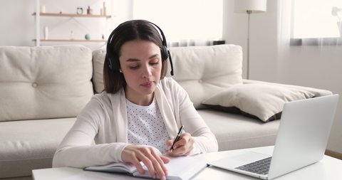 Young girl student wearing wireless headset e learning by web cam chat at home. Millennial woman video conference calling on laptop computer making notes talking by webcam. Distance education concept