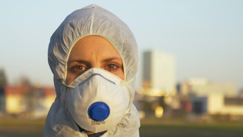 SLOW MOTION, CLOSE UP, PORTRAIT, DOF: Young nurse wearing a protective facemask and suit stands at a coronavirus safety checkpoint on sunny morning. Caucasian nurse during the covid-19 outbreak.