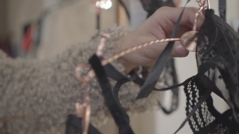 Close-up of female young hands touching lace underwear on hanger. Caucasian wealthy woman choosing undergarment in fashion boutique. Elegance, lifestyle. Slow motion.