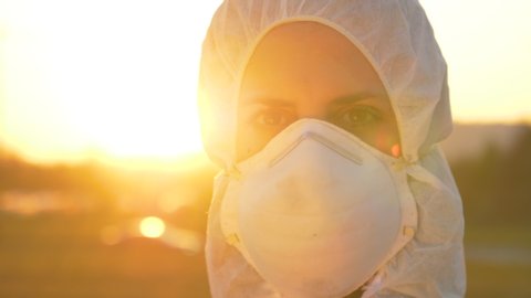 SLOW MOTION, CLOSE UP, LENS FLARE, PORTRAIT, DOF: Female doctor wearing a facemask and suit stands outside in the golden sunset. Nurse wears a protective suit and mask during the COVID19 outbreak