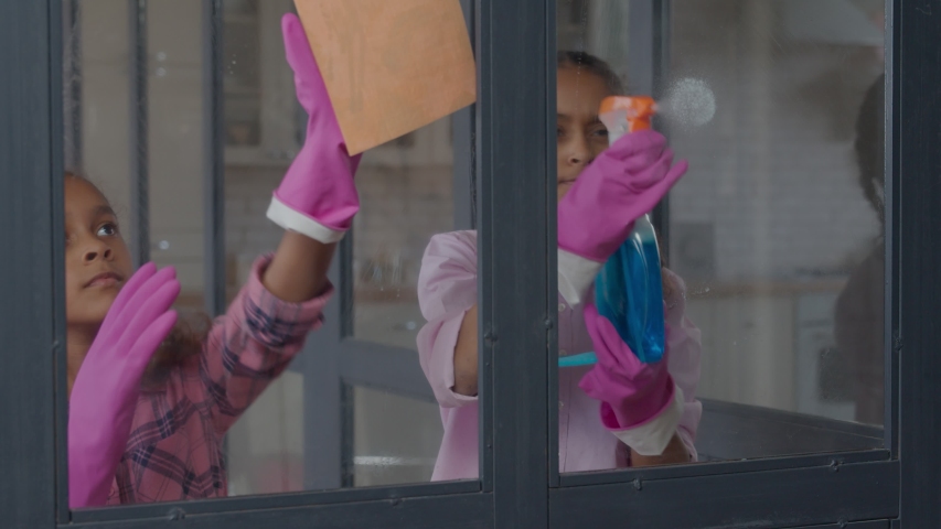 Lovely engrossed in household chores preadolescent african american girls in rubber gloves cleaning windows glass with rags and detergent spray during spring cleanup, looking enthusiastic and happy,