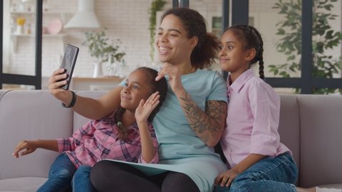 Positive united african american family with two cute elementary age daughters making video call online using cellphone while siiting on sofa at home. Mom and girls with smart phone video conferencing