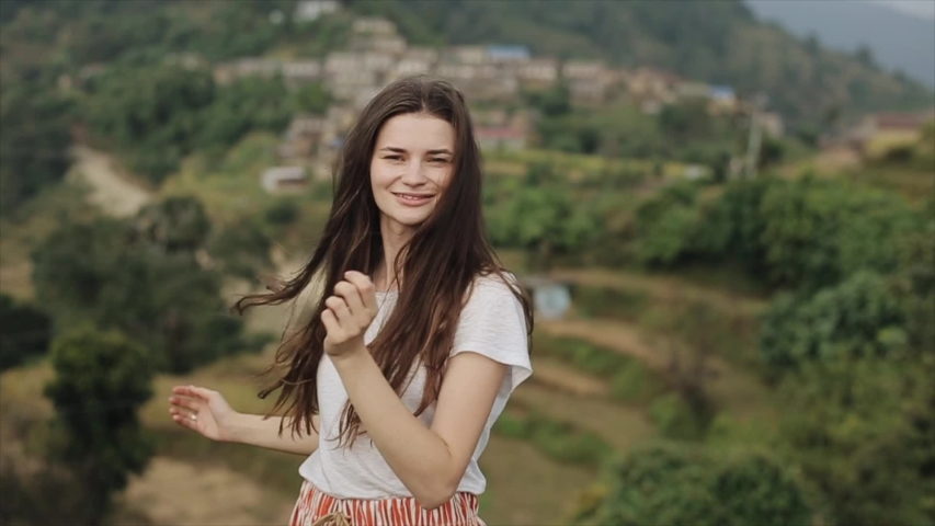 A young slim caucasian girl with long brown hair spinning and smiling at camera on a top of a mountain, hill in Nepal. Landscape