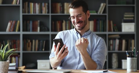 Amazed happy businessman receiving sms message reading good news. Excited overjoyed male winner celebrating success looking at smart phone sitting at home office desk. Mobile victory concept