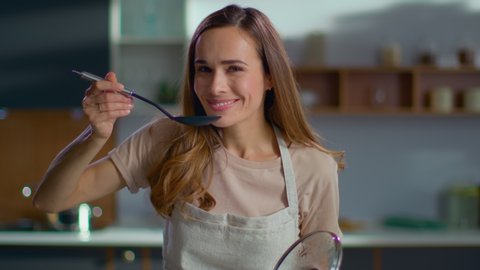 Pretty woman cooking soup on stove at kitchen. Close up of housewife preparing dinner at home. Young woman tasting soup on spoon at modern kitchen in slow motion