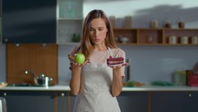 Cheerful woman making choice between healthy and unhealthy food on kitchen. Girl looking on apple and cake on plates at home in slow motion. Young woman choosing green apple instead cake on kitchen