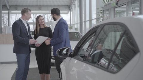 Young rich multiracial family buying car in dealership. Smiling Middle Eastern client and Caucasian dealer shaking hands in showroom. Agreement, business, luxury, success.