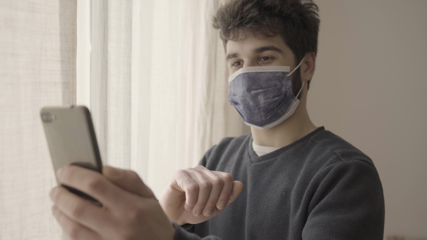 Young positive man regarding family wearing protective mask on a phone video call in hospital, standing by a window, coronavirus isolation, bye bye gesture, blurred final.  | Shutterstock HD Video #1048500808