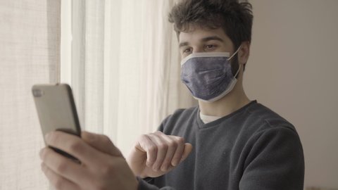 Young positive man regarding family wearing protective mask on a phone video call in hospital, standing by a window, coronavirus isolation, bye bye gesture, blurred final. 