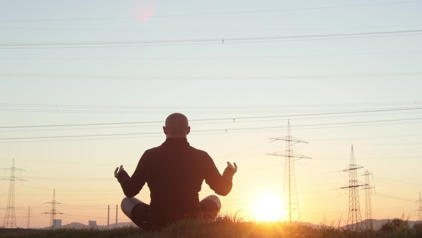 Yoga time. Young bald bearded man in casual wear relaxing in green field after work, meditation of male against sunset or sunrise.