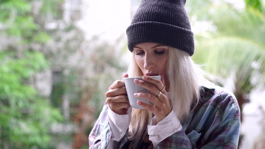 blond hipster girl in black har and plaid shirt is sick and happy drinking hot tea or coffee and smiling on camera on sunny and gloomy day palm trees in 4k