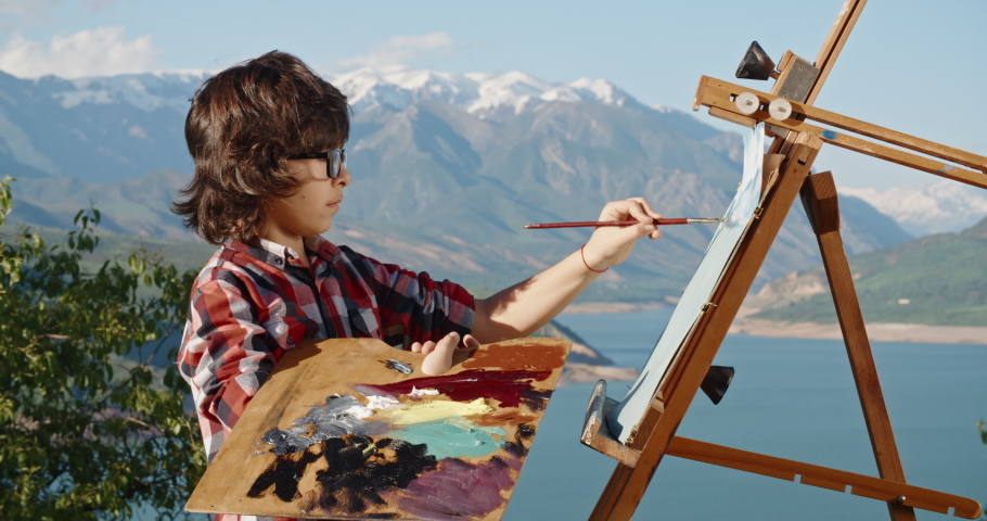 Young kid standing in front of easel, learning how to draw, creating a picture inspired by beautiful landscape, making first steps in fulfilling childhood dream - childhood memories, education concept