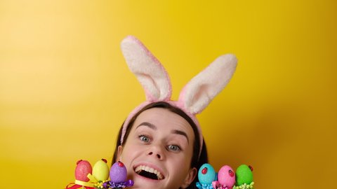 Charming cheerful happy woman sits at desk near colored Easter eggs, wears fluffy bunny ears, fun looks and hides face, models over yellow background. Spring holiday and Easter concept.