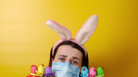 Displeased young woman with virus mask, wears fluffy ears, sad to celebrate Easter alone, holds colored eggs on yellow background with copy space. Coronavirus outbreak Holiday concept.