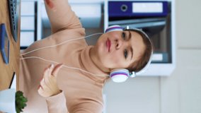 Happy female worker in headphones listening to music in office. Vertical video 