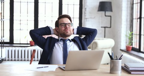 Happy relaxed businessman wears suit finished project on computer stretching at workplace. Smiling ceo feels stress relief after work well done takes break sits at office desk puts hands behind head.