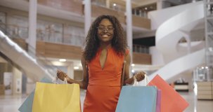 Fashion girl doing shopping in mall center. Happy afro-american woman having fun buying new clothes. Excited girl raising hands with shopping bags demonstrating purchases and smiling