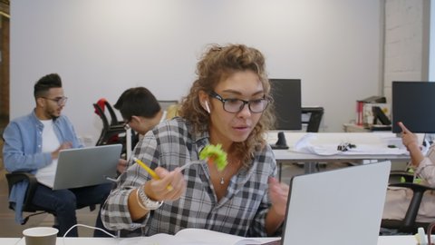Young mixed raced woman in wireless headphones and casualwear eating vegetable salad, speaking via video call on laptop and taking notes while working in coworking space