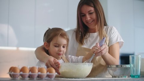 Caucasian mother and daughter having fun while making dough on kitchen. Happy family in the kitchen. Child daughter preparing the dough, bake cookies.